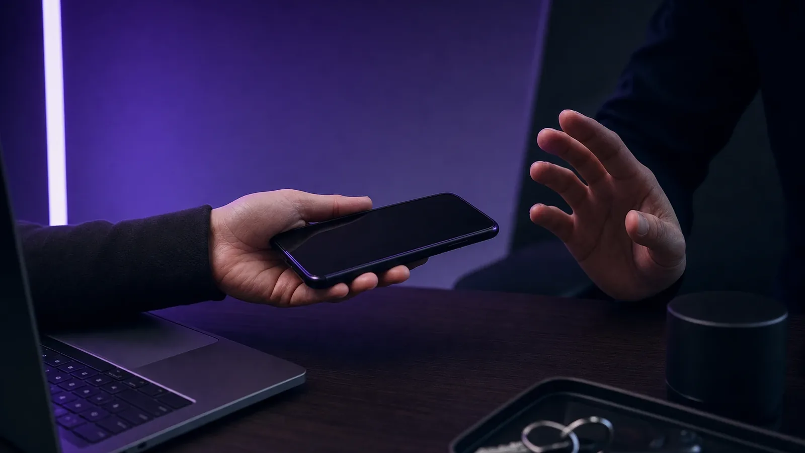 Una mano ofrece un móvil a otra que duda, sobre una mesa oscura con un portátil y unas llaves